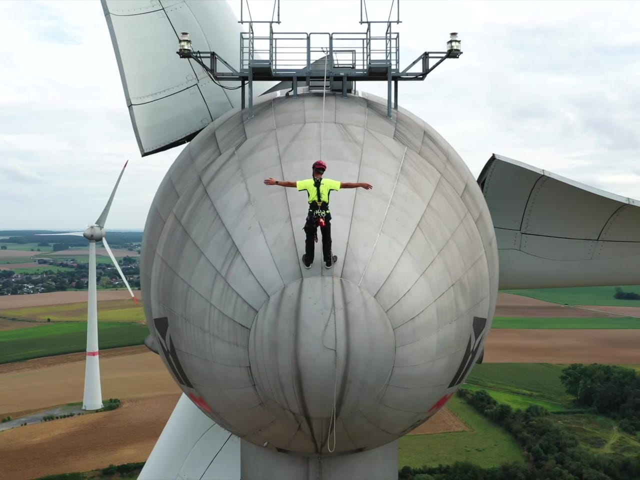 Technicien bras écartés sur la nacelle d'une éolienne, filmé par drone